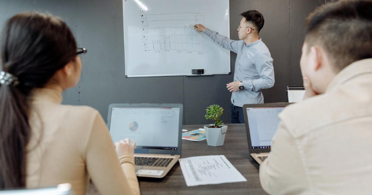 Person at whiteboard conducting sales goals meeting before people with laptops